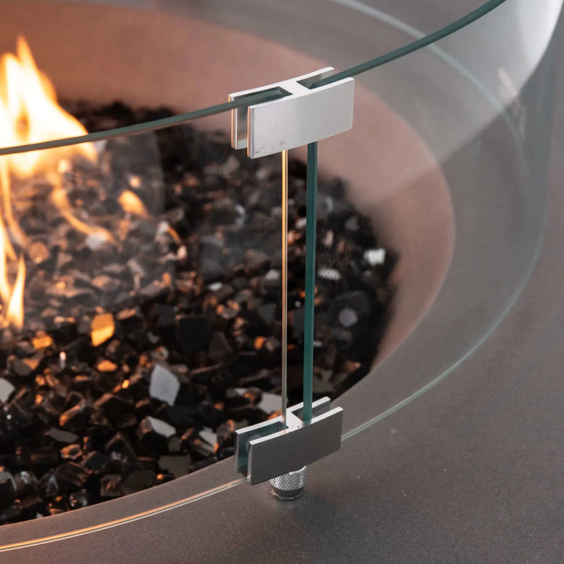 Close-up of the tempered glass wind guard mounting hardware on the Flame Havens Refuge Series 43-inch Round Propane Gas GFRC Concrete Fire Pit in Gray. The image highlights the precision stainless steel glass brackets, premium tempered glass panels, and black reflective fire glass surrounding the propane flame, showcasing the quality engineering and secure wind guard installation.