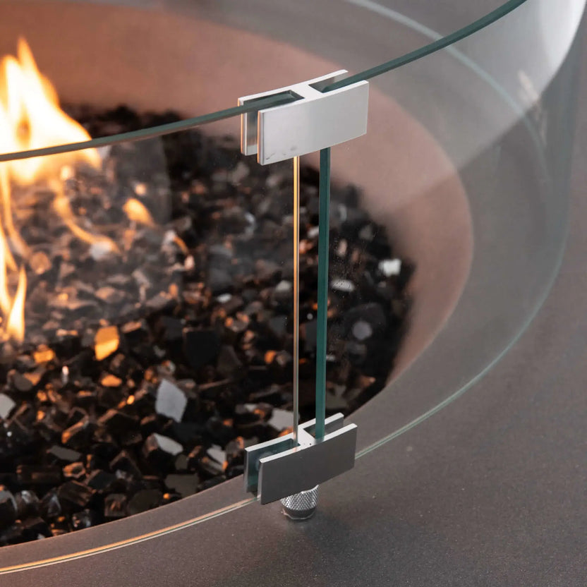 Close-up of the tempered glass wind guard mounting hardware on the Flame Havens Refuge Series 43-inch Round Propane Gas GFRC Concrete Fire Pit in Gray. The image highlights the precision stainless steel glass brackets, premium tempered glass panels, and black reflective fire glass surrounding the propane flame, showcasing the quality engineering and secure wind guard installation.