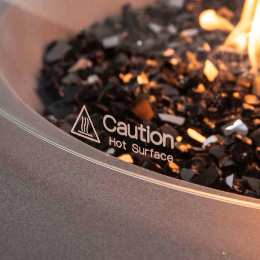 Detailed close-up of the tempered glass wind guard on the Flame Havens Refuge Series 43-inch Round Propane Gas GFRC Concrete Fire Pit in Gray. The image focuses on the etched "Caution Hot Surface" safety marking, tempered black reflective fire glass, and active propane flame, illustrating the premium safety features and refined finish of the optional glass wind guard accessory.