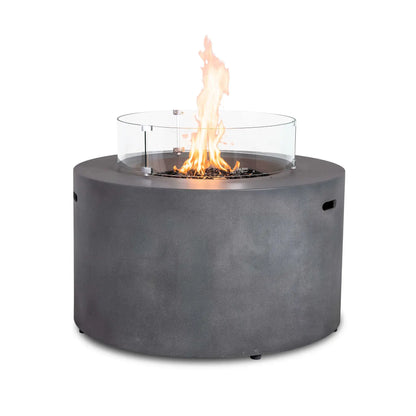 The Flame Havens Refuge Series 43-inch Round Propane Gas GFRC Concrete Fire Pit in Gray featuring premium tempered black reflective fire glass and the optional tempered glass wind guard. The tall flame is protected by the clear circular wind guard, providing enhanced flame stability while preserving unobstructed views of the modern round fire pit.