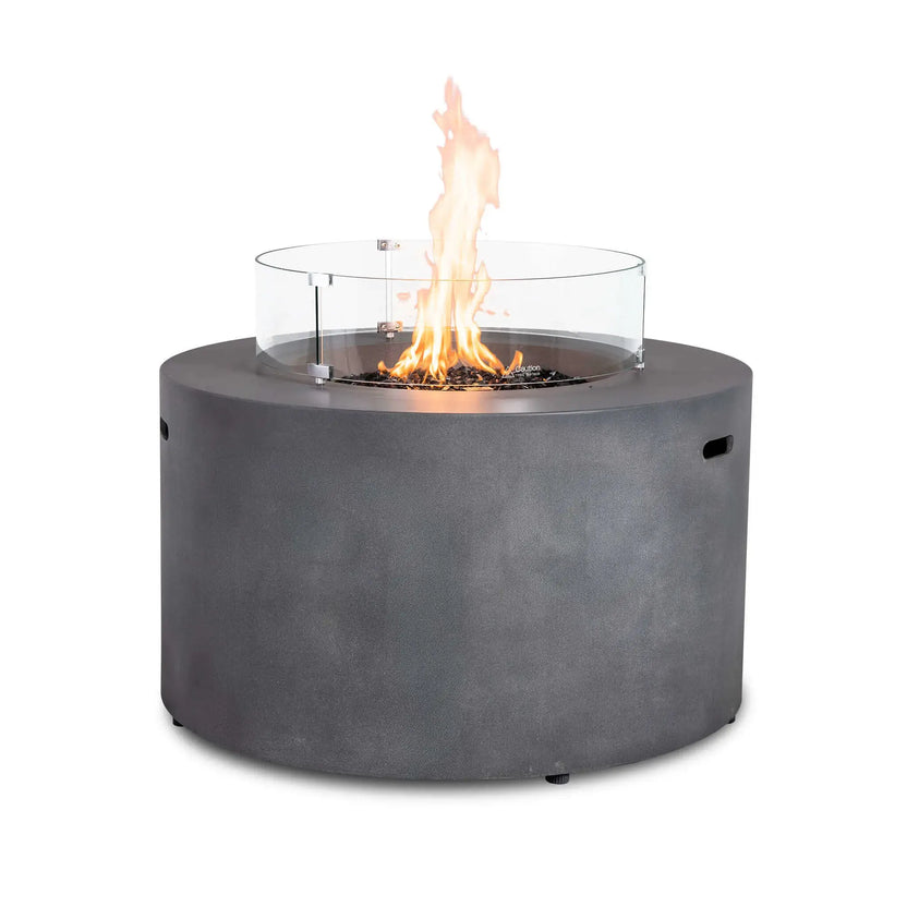 The Flame Havens Refuge Series 43-inch Round Propane Gas GFRC Concrete Fire Pit in Gray featuring premium tempered black reflective fire glass and the optional tempered glass wind guard. The tall flame is protected by the clear circular wind guard, providing enhanced flame stability while preserving unobstructed views of the modern round fire pit.