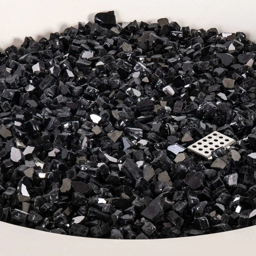 Detailed close-up of the tempered black reflective fire glass inside the Flame Havens Refuge Series 43-inch Round Propane Gas GFRC Concrete Fire Pit in White. The image highlights the premium reflective glass media surrounding the stainless steel CSA ignition component, showcasing the quality, depth, and polished finish of the fire feature.
