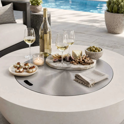 Lifestyle image of the Flame Havens Refuge Series 43-inch Round Propane Gas GFRC Concrete Fire Pit in White with the optional stainless steel burner cover installed as a tabletop. Styled with a wine bottle, glasses, charcuterie board, appetizers, olives, and candle beside a luxury pool, the image demonstrates how the fire pit doubles as an elegant entertaining table when the burner is covered.