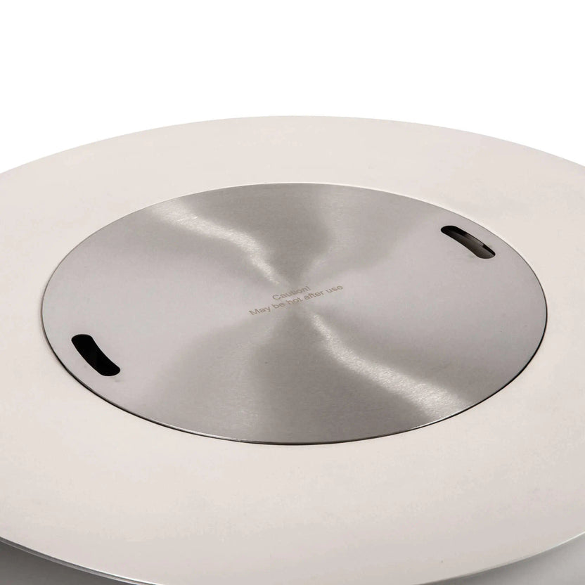 The Flame Havens Refuge Series 43-inch Round Propane Gas GFRC Concrete Fire Pit in White fitted with the optional brushed stainless steel burner cover. The cover sits flush within the burner opening, transforming the fire pit into a stylish outdoor table while preserving the clean lines and premium GFRC concrete construction.