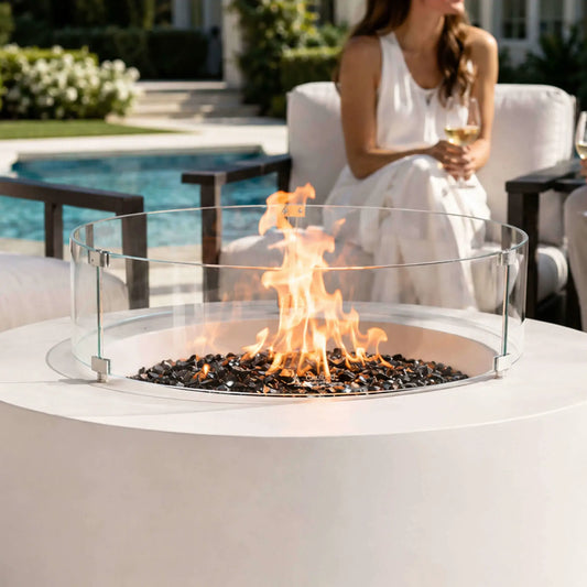 Lifestyle image of the Flame Havens Refuge Series 43-inch Round Propane Gas GFRC Concrete Fire Pit in White featuring premium tempered black reflective fire glass and the optional tempered glass wind guard. Positioned beside a luxury swimming pool with modern outdoor seating, the fire pit creates a warm focal point for elegant outdoor entertaining.