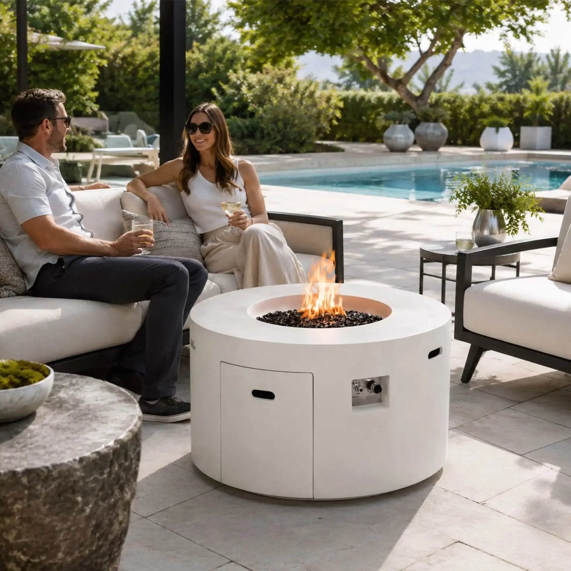Lifestyle image of the Flame Havens Refuge Series 43-inch Round Propane Gas GFRC Concrete Fire Pit in White burning with premium tempered black reflective fire glass in a luxurious backyard seating area. A couple relaxes beside the fire pit overlooking a swimming pool, showcasing how the Refuge Series creates an inviting centerpiece for outdoor entertaining and modern patio living.