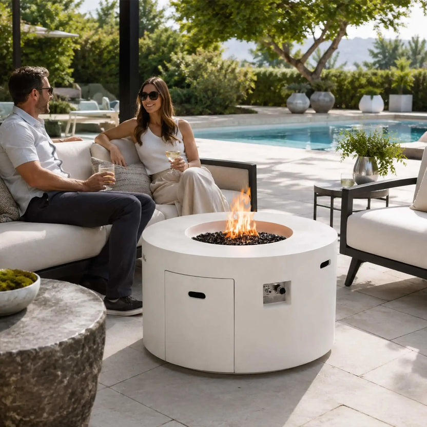 Lifestyle image of the Flame Havens Refuge Series 43-inch Round Propane Gas GFRC Concrete Fire Pit in White burning with premium tempered black reflective fire glass in a luxurious backyard seating area. A couple relaxes beside the fire pit overlooking a swimming pool, showcasing how the Refuge Series creates an inviting centerpiece for outdoor entertaining and modern patio living.