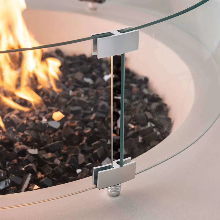 Close-up of the tempered glass wind guard mounting hardware on the Flame Havens Refuge Series 43-inch Round Propane Gas GFRC Concrete Fire Pit in White. The image highlights the precision stainless steel brackets, crystal-clear tempered glass panels, premium tempered black reflective fire glass, and active flame, demonstrating the quality craftsmanship and secure wind guard installation.