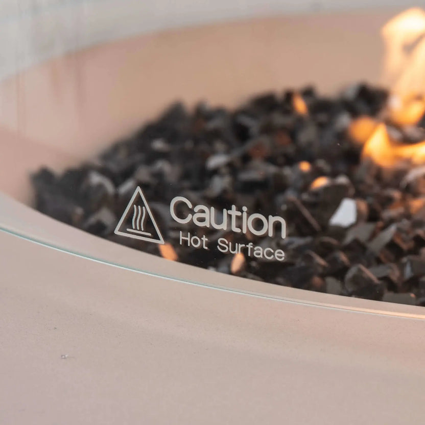Detailed close-up of the etched "Caution Hot Surface" marking on the optional tempered glass wind guard installed on the Flame Havens Refuge Series 43-inch Round Propane Gas GFRC Concrete Fire Pit in White. Premium tempered black reflective fire glass and natural flames remain visible behind the clear glass, emphasizing both safety and the refined design of this luxury outdoor fire feature.