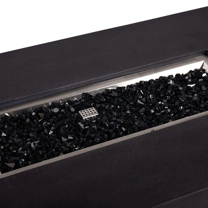 Close-up angled view of a Flame Havens Refuge Series 56 x 30 x 15 inch rectangle natural gas GFRC concrete fire pit in black, showing the stainless steel burner pan filled with glossy black fire glass and the CSA-style ignition cage centered in the media bed.