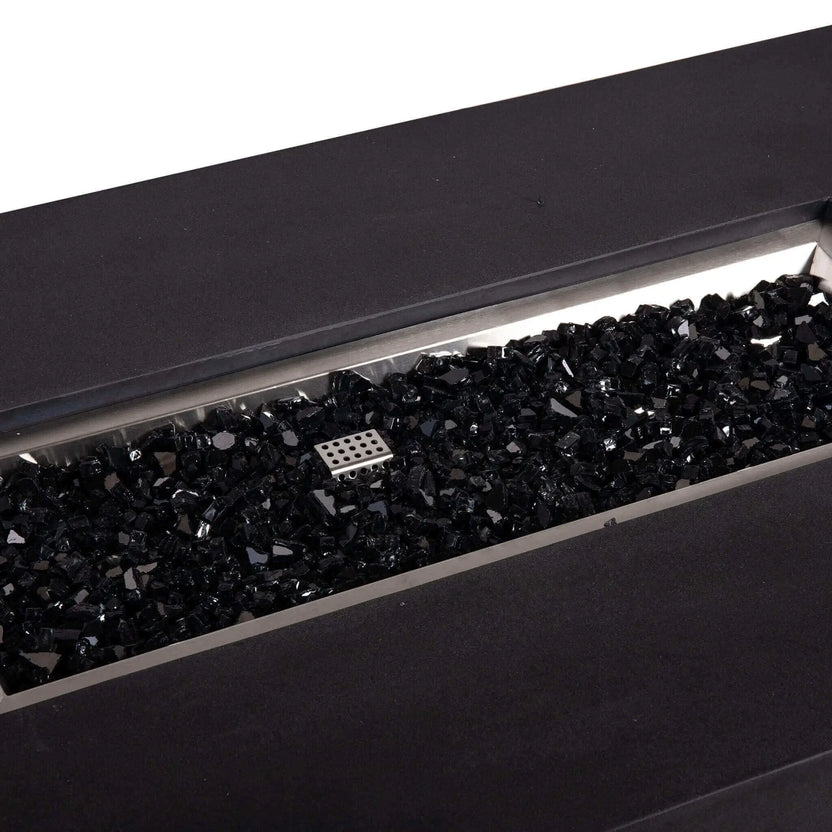 Close-up angled view of a Flame Havens Refuge Series 56 x 30 x 15 inch rectangle natural gas GFRC concrete fire pit in black, showing the stainless steel burner pan filled with glossy black fire glass and the CSA-style ignition cage centered in the media bed.