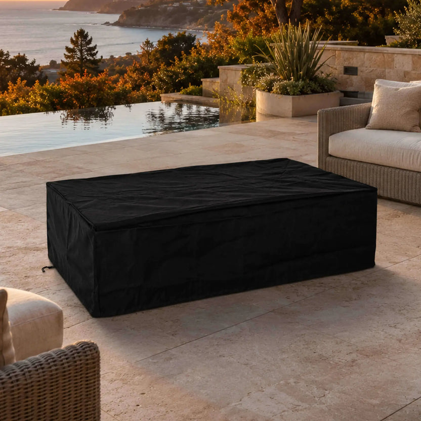 Flame Havens Refuge Series 56 x 30 x 15 inch rectangle natural gas black GFRC concrete fire pit covered with a fitted black canvas fabric cover in a luxury outdoor patio setting overlooking an infinity pool and coastline at sunset.