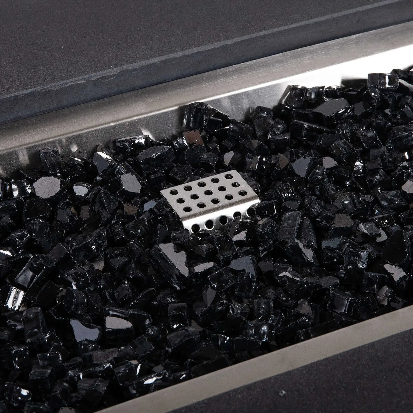 Detailed close-up of the Flame Havens Refuge Series 56 x 30 x 15 inch rectangle angled natural gas fire pit in black, featuring reflective black fire glass surrounding the stainless steel ignition cage inside the brushed stainless burner pan.
