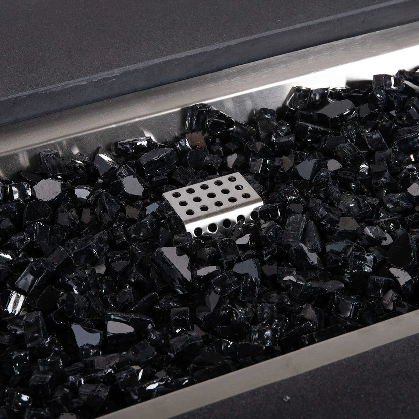 Detailed close-up of the Flame Havens Refuge Series 56 x 30 x 15 inch rectangle angled natural gas fire pit in black, featuring reflective black fire glass surrounding the stainless steel ignition cage inside the brushed stainless burner pan.