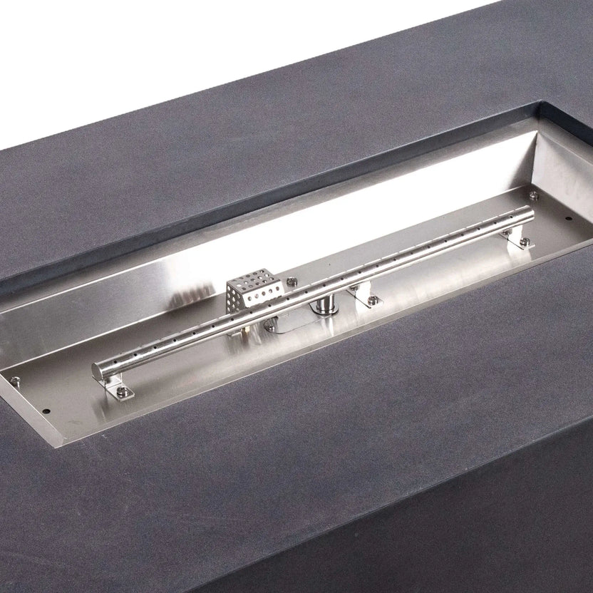 Tight close-up of the Flame Havens Refuge Series 56 x 30 x 15 inch rectangle natural gas fire pit burner system, showing the stainless steel linear burner, CSA ignition cage, mounting hardware, and brushed metal pan set into the black GFRC concrete top.