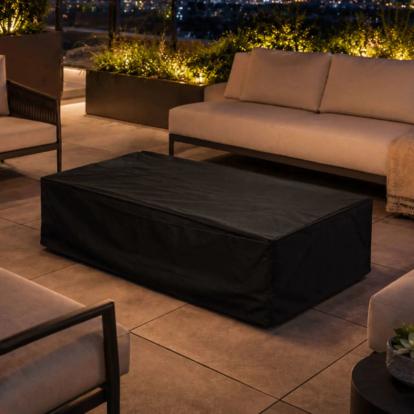 Lifestyle image of the Flame Havens Refuge Series 56 x 30 x 15 inch rectangle natural gas gray GFRC fire pit protected with its custom black canvas fabric cover. Positioned within a luxury outdoor seating area at night, the covered fire pit blends seamlessly into a sophisticated patio designed for year-round protection.