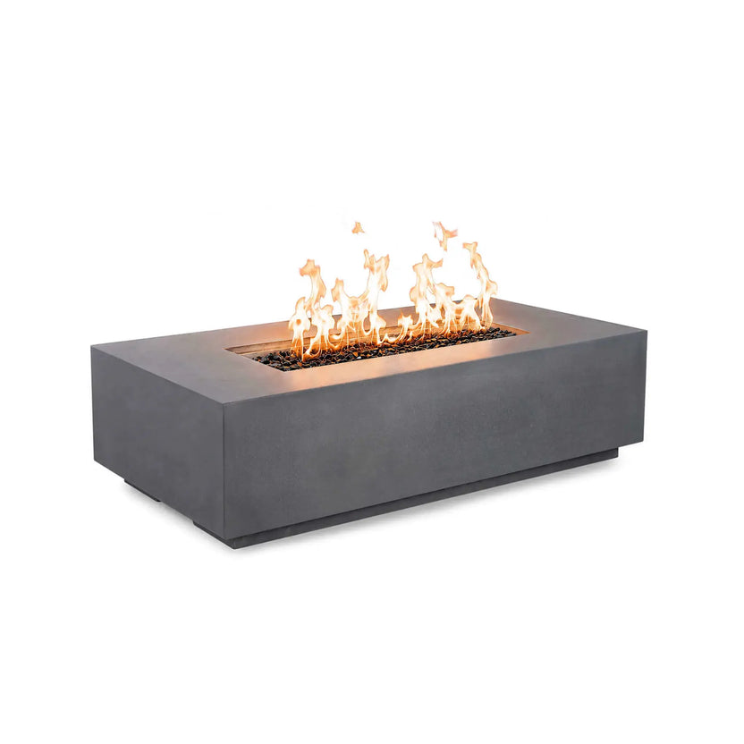 Angled front view of the Flame Havens Refuge Series 56 x 30 x 15 inch rectangle natural gas gray GFRC fire pit burning with bright flames over reflective tempered black fire glass. The streamlined design features concealed controls, creating a clean, contemporary appearance.