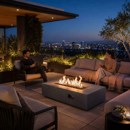 Lifestyle scene featuring the Flame Havens Refuge Series 56 x 30 x 15 inch rectangle natural gas gray GFRC fire pit with reflective tempered black fire glass burning brightly on a luxury rooftop patio. Friends relax around the fire while enjoying panoramic city views, demonstrating the fire pit as the centerpiece of an upscale outdoor living space.