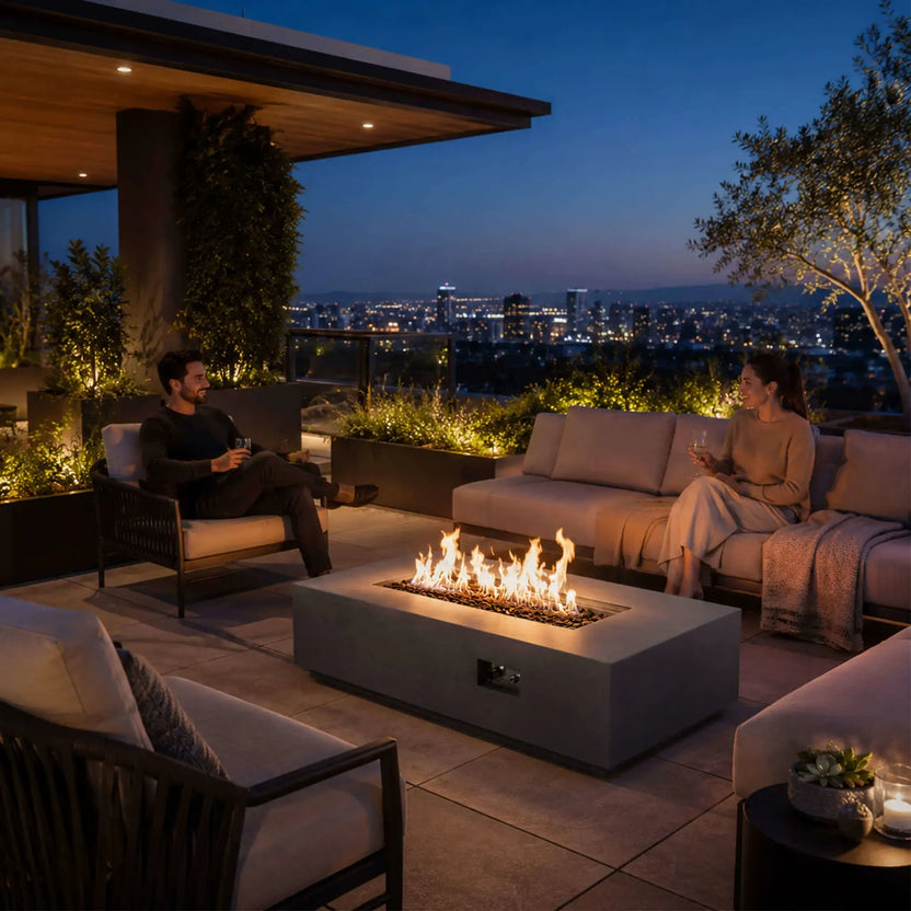 Lifestyle scene featuring the Flame Havens Refuge Series 56 x 30 x 15 inch rectangle natural gas gray GFRC fire pit with reflective tempered black fire glass burning brightly on a luxury rooftop patio. Friends relax around the fire while enjoying panoramic city views, demonstrating the fire pit as the centerpiece of an upscale outdoor living space.