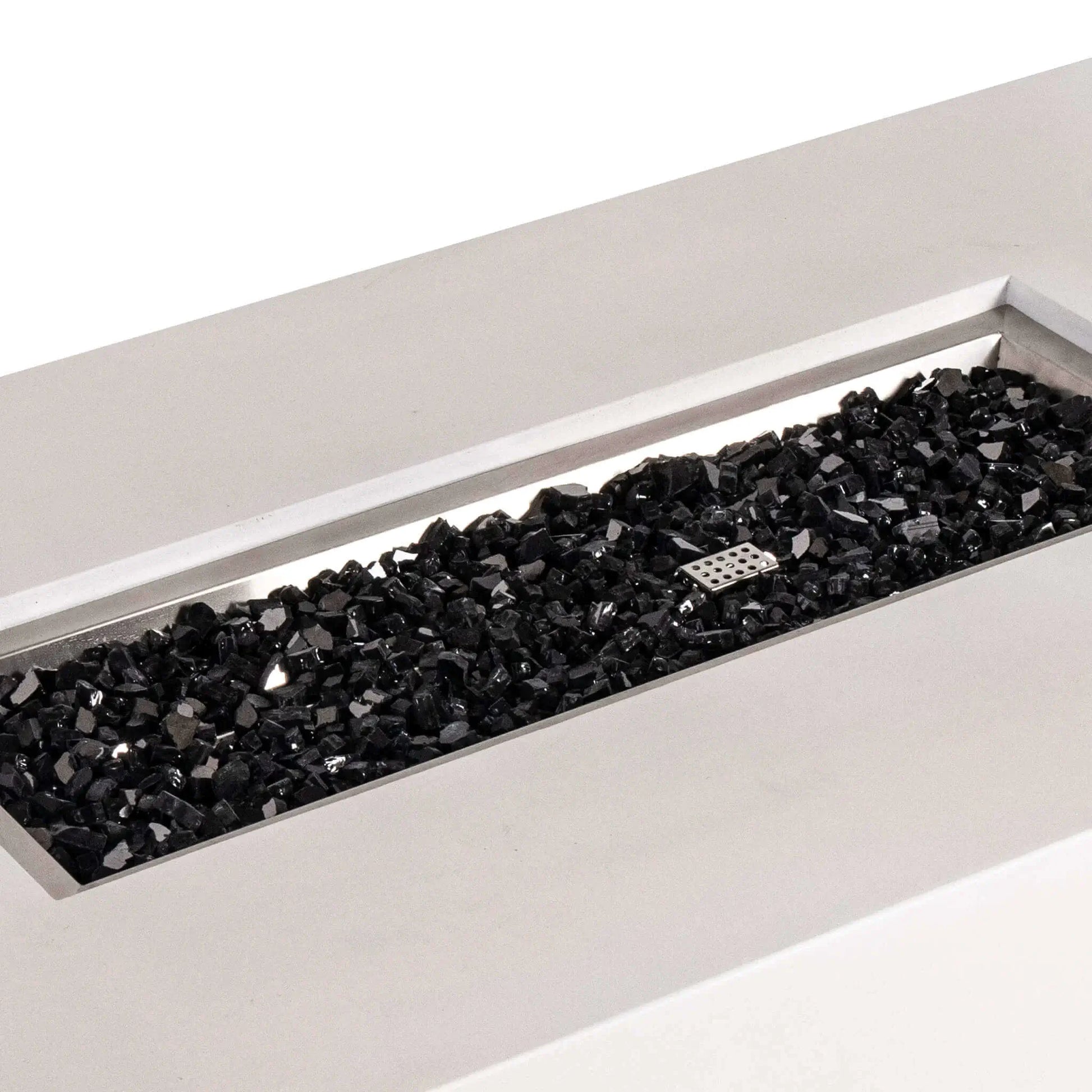 Angled close-up of the Flame Havens Refuge Series 56 x 30 x 15 inch rectangle natural gas white GFRC concrete fire pit showcasing reflective black fire glass neatly filling the brushed stainless steel burner pan. The image highlights the premium fire media, clean rectangular opening, and modern craftsmanship designed for luxury outdoor living.