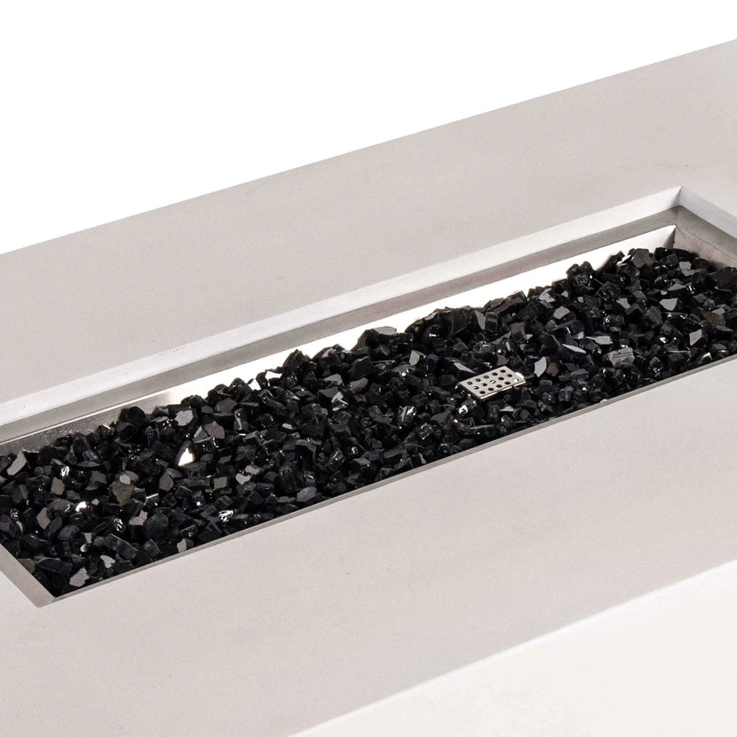 Angled close-up of the Flame Havens Refuge Series 56 x 30 x 15 inch rectangle natural gas white GFRC concrete fire pit showcasing reflective black fire glass neatly filling the brushed stainless steel burner pan. The image highlights the premium fire media, clean rectangular opening, and modern craftsmanship designed for luxury outdoor living.