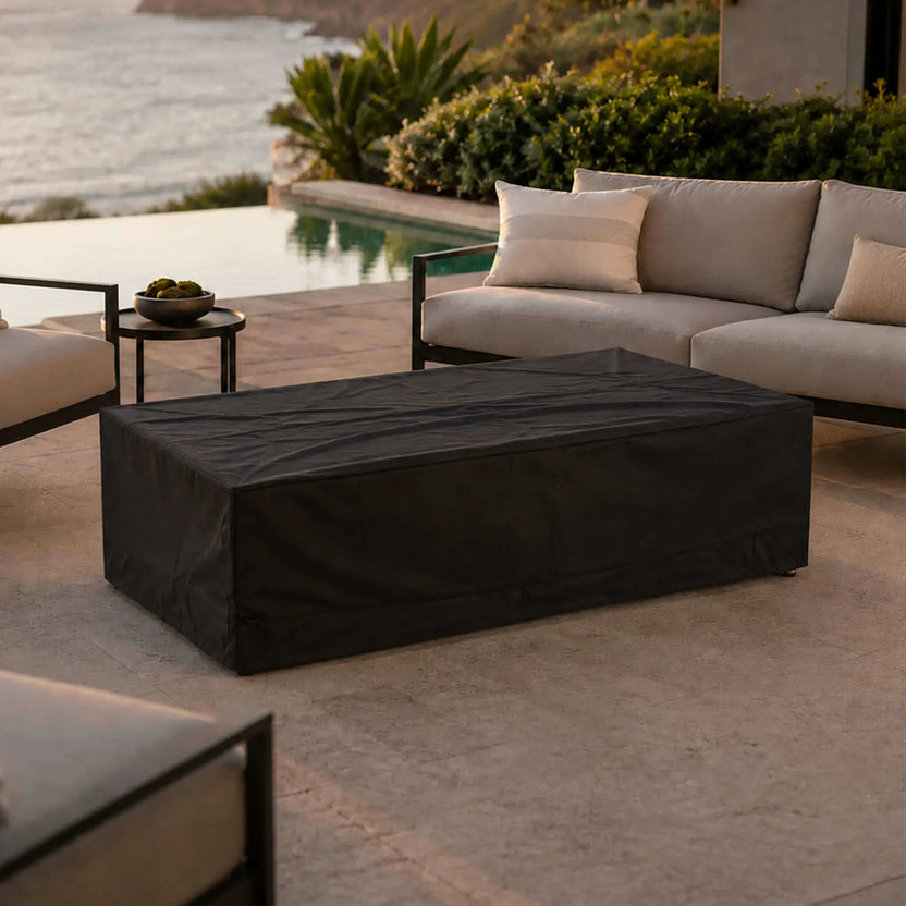 Lifestyle view of the Flame Havens Refuge Series 56 x 30 x 15 inch rectangle natural gas white GFRC concrete fire pit protected by its black canvas fabric cover. Positioned between contemporary outdoor seating beside an infinity pool overlooking the coastline, this image demonstrates secure off-season protection while complementing an upscale outdoor entertaining space.