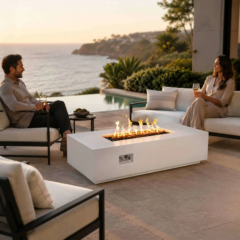 Lifestyle image of the Flame Havens Refuge Series 56 x 30 x 15 inch rectangle natural gas white GFRC concrete fire pit burning brightly on a luxury coastal patio. Two people relax with drinks beside the fire while reflective tempered black fire glass and the recessed control panel complement the sophisticated outdoor entertaining space.