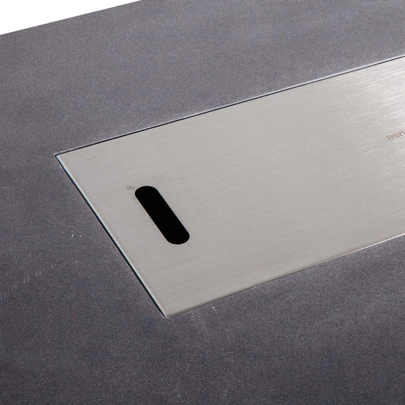 Close-up of the Flame Havens Refuge Series 56 x 30 x 15 inch rectangle natural gas black GFRC fire pit with a flush stainless steel burner cover installed, showing the brushed metal surface, finger pull opening, and clean concrete top finish.