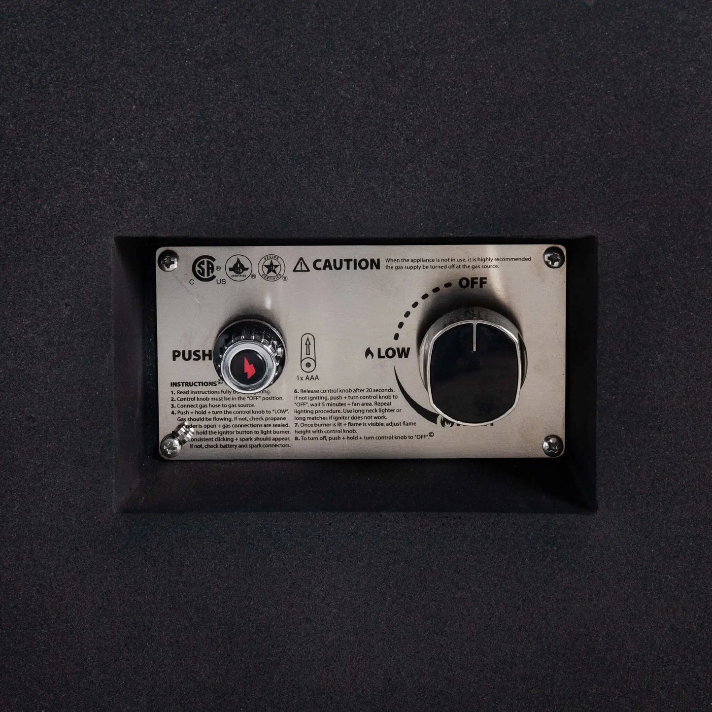 Detailed close-up of the Flame Havens Refuge Series 56 x 30 x 15 inch rectangle natural gas fire pit control panel, highlighting the CSA-certified ignition system, push-button spark igniter, adjustable gas control knob, and stainless steel mounting plate recessed into the black GFRC body.