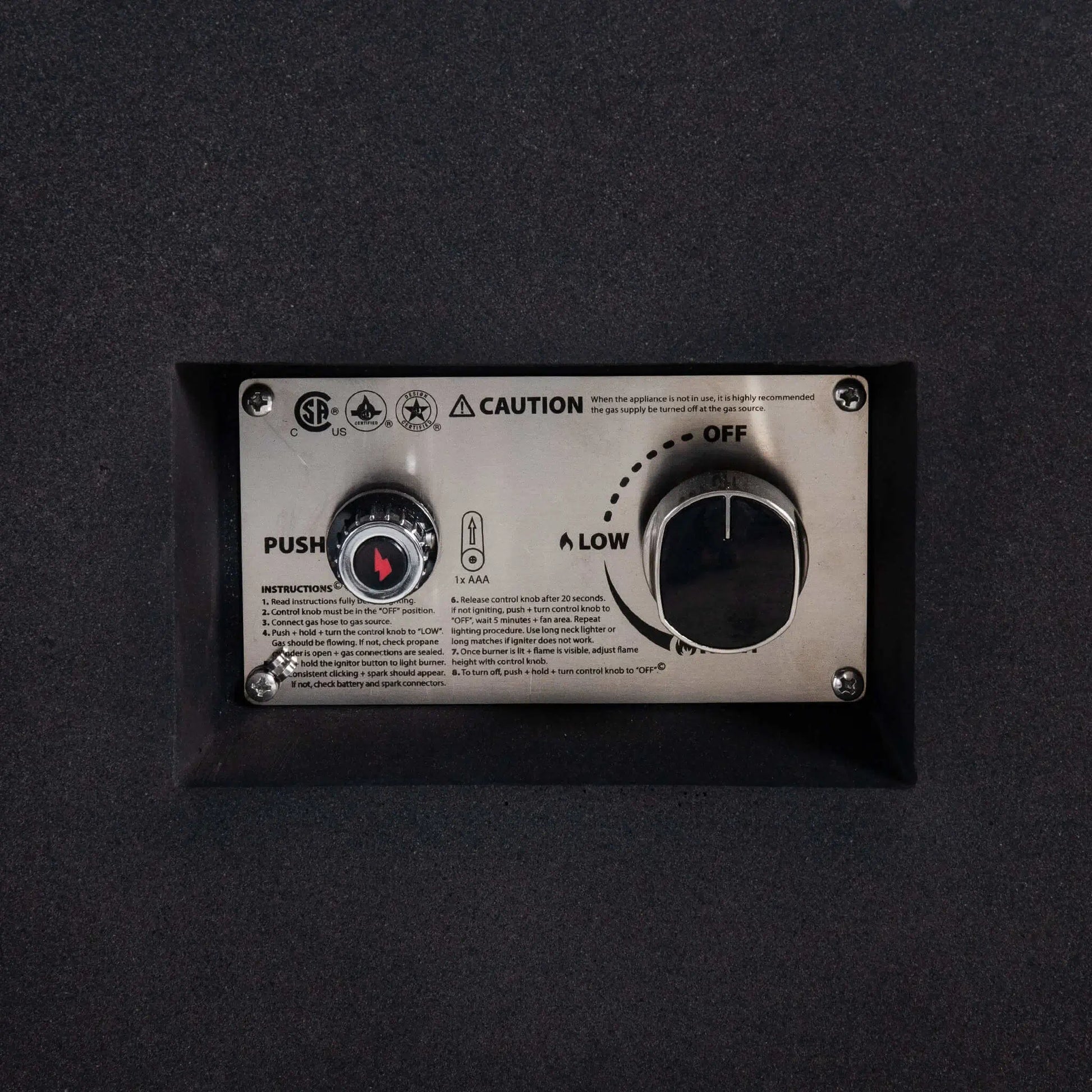 Detailed close-up of the Flame Havens Refuge Series 56 x 30 x 15 inch rectangle natural gas fire pit control panel, highlighting the CSA-certified ignition system, push-button spark igniter, adjustable gas control knob, and stainless steel mounting plate recessed into the black GFRC body.