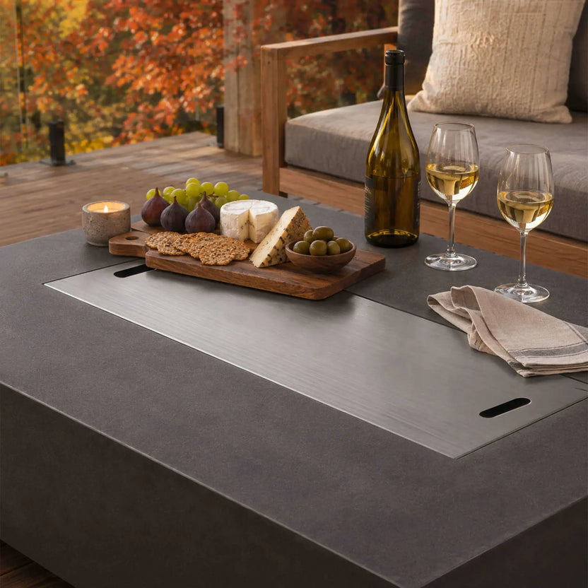 Flame Havens Refuge Series 56 x 30 x 15 inch rectangle natural gas black GFRC fire pit with the stainless steel burner cover installed and used as an outdoor entertaining table, displaying a charcuterie board, wine, and appetizers on a cozy autumn patio.