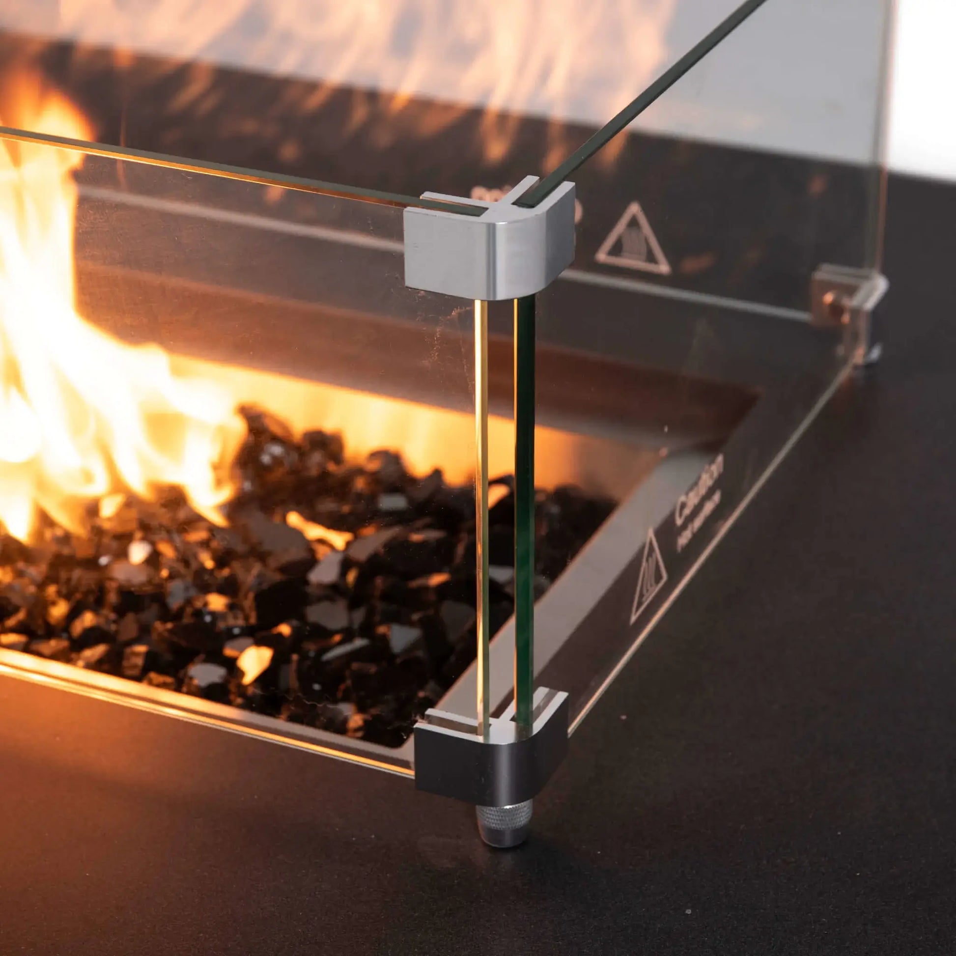 Close-up of the Flame Havens Refuge Series rectangle natural gas fire pit with the tempered glass wind guard installed, showcasing polished corner brackets, black reflective fire glass, and vibrant flames contained behind the premium glass panels.