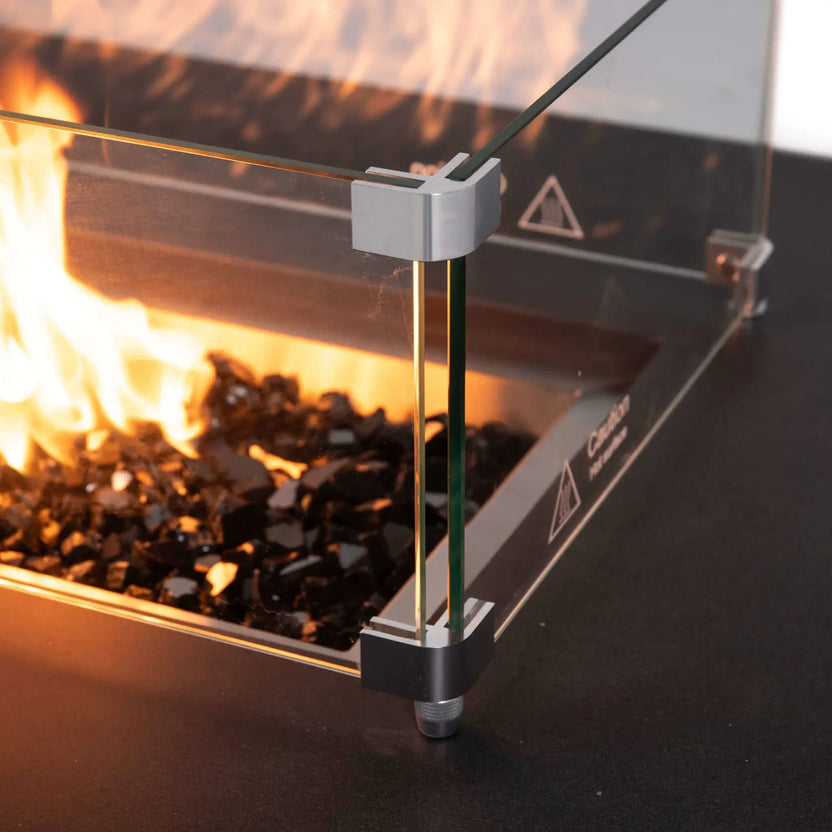 Close-up of the Flame Havens Refuge Series rectangle natural gas fire pit with the tempered glass wind guard installed, showcasing polished corner brackets, black reflective fire glass, and vibrant flames contained behind the premium glass panels.