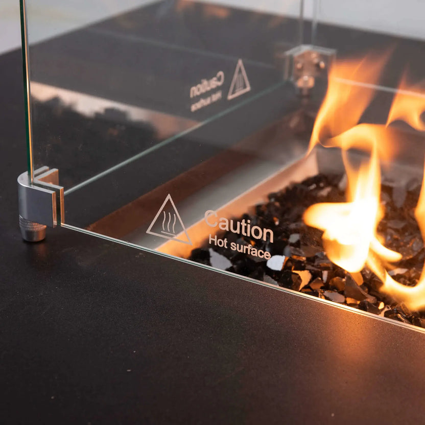 Detailed view of the Flame Havens Refuge Series tempered glass wind guard mounted on the 56 x 30 x 15 inch rectangle natural gas fire pit, featuring etched caution markings, polished hardware, black fire glass, and warm flames behind the safety glass.
