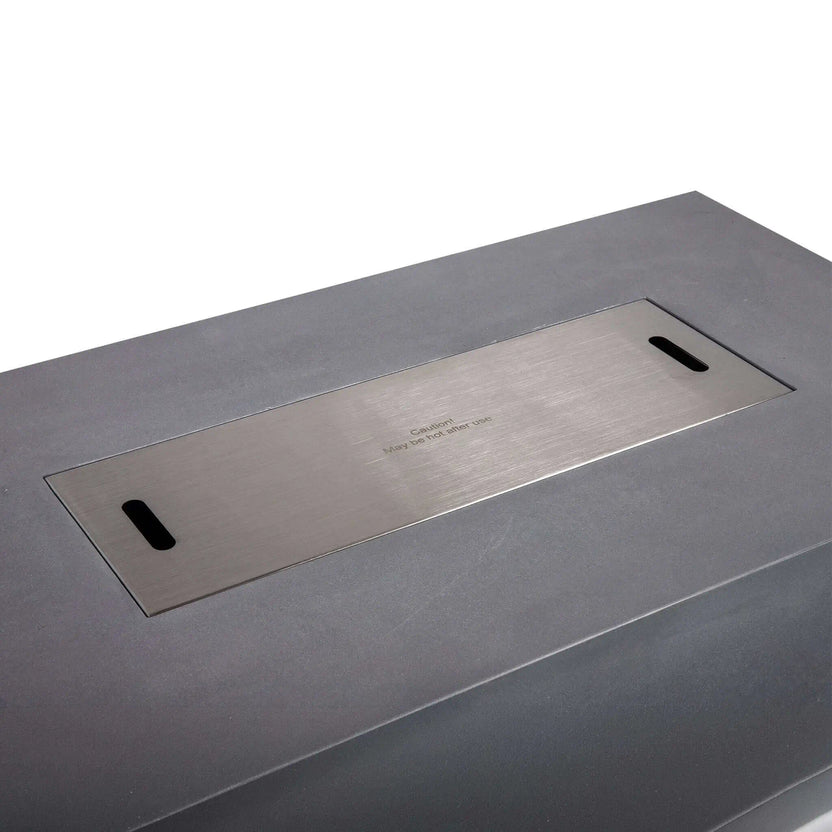 Angled close-up of the Flame Havens Refuge Series 56 x 30 x 15 inch rectangle natural gas gray GFRC concrete fire pit with the brushed stainless steel burner cover installed. The precision-fit cover sits flush within the burner opening and features integrated lift slots, creating a clean tabletop surface when the fire pit is not in use.
