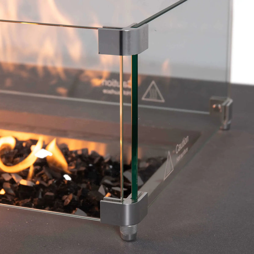 Close-up of the Flame Havens Refuge Series 56 x 30 x 15 inch rectangle natural gas gray GFRC fire pit featuring the tempered glass wind guard with brushed stainless steel corner brackets. Flames rise above reflective tempered black fire glass while the wind guard provides added protection without obstructing the fire.
