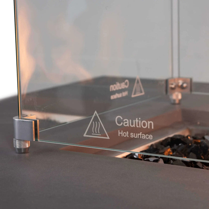 Detailed view of the Flame Havens Refuge Series 56 x 30 x 15 inch rectangle natural gas gray fire pit with tempered glass wind guard installed. The etched caution markings, stainless steel mounting hardware, and reflective tempered black fire glass demonstrate the premium craftsmanship and safety-focused design.