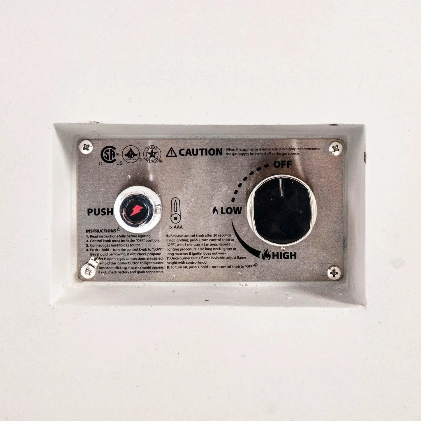 Detailed close-up of the Flame Havens Refuge Series 56 x 30 x 15 inch rectangle natural gas white GFRC concrete fire pit showing the recessed CSA-certified ignition control panel. The stainless steel faceplate features the push-button igniter, flame adjustment knob, operating instructions, and safety markings for reliable natural gas operation.