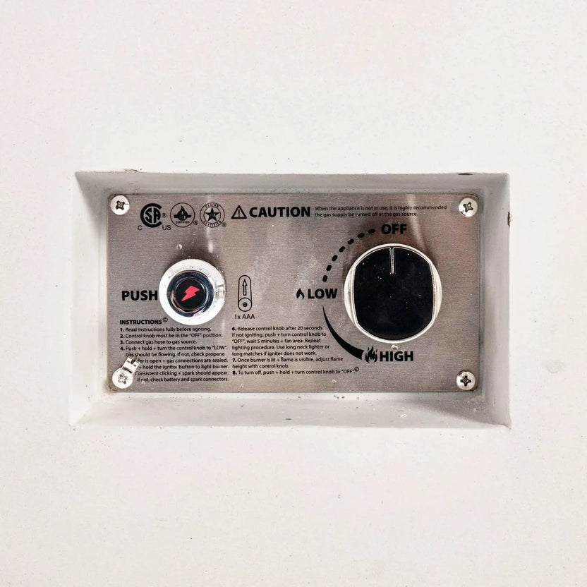 Detailed close-up of the Flame Havens Refuge Series 56 x 30 x 15 inch rectangle natural gas white GFRC concrete fire pit showing the recessed CSA-certified ignition control panel. The stainless steel faceplate features the push-button igniter, flame adjustment knob, operating instructions, and safety markings for reliable natural gas operation.