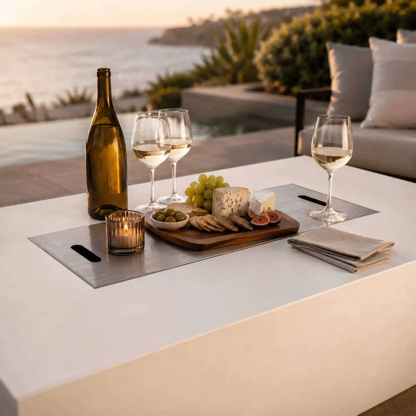 Lifestyle image of the Flame Havens Refuge Series 56 x 30 x 15 inch rectangle natural gas white GFRC concrete fire pit with the stainless steel burner cover installed as a tabletop. A wine bottle, glasses, cheese board, grapes, crackers, and candle create an inviting outdoor entertaining space overlooking an infinity pool and ocean at sunset.
