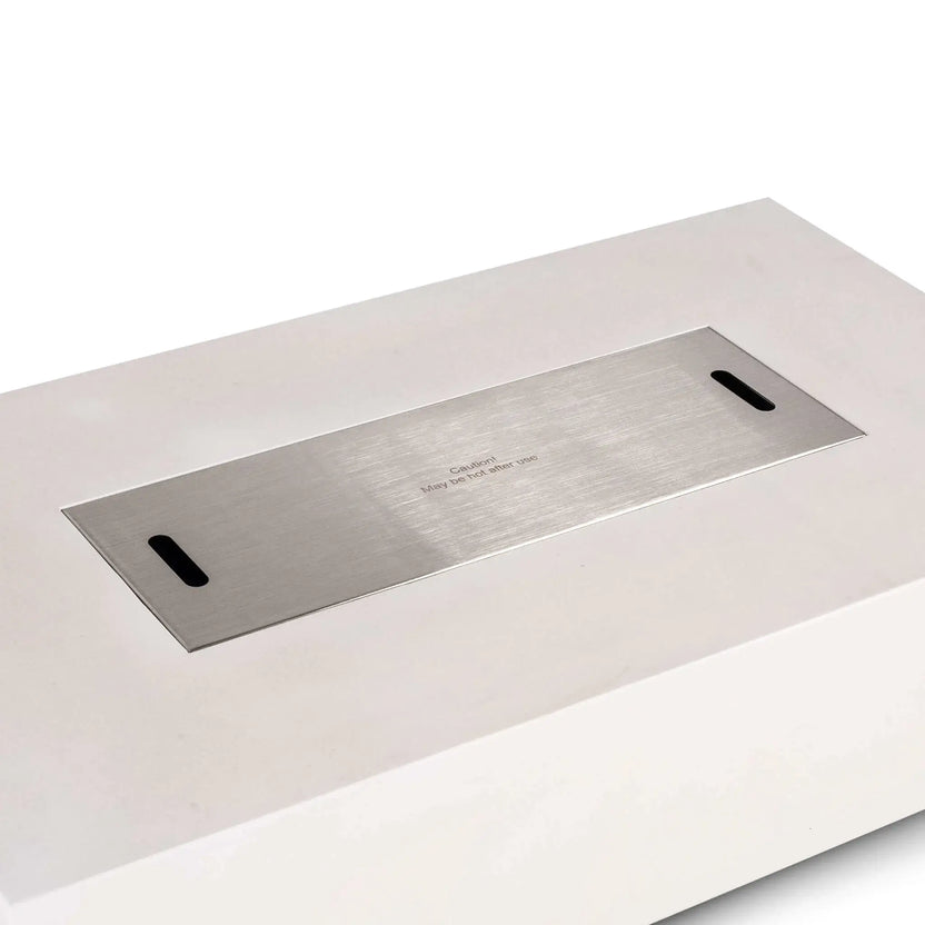Close-up of the Flame Havens Refuge Series 56 x 30 x 15 inch rectangle natural gas white GFRC concrete fire pit featuring the brushed stainless steel burner cover installed flush within the burner opening. Dual lift slots and the laser-etched caution marking highlight the premium finish and protective cover designed to transform the fire pit into a functional tabletop when not in use.