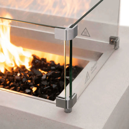 Close-up of the Flame Havens Refuge Series 56 x 30 x 15 inch rectangle natural gas white GFRC concrete fire pit featuring the tempered glass wind guard surrounding lively flames over reflective tempered black fire glass. Stainless steel corner brackets securely support the glass while preserving clear views of the fire.