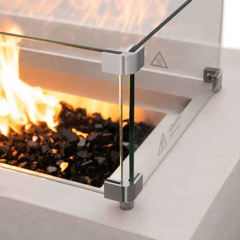 Close-up of the Flame Havens Refuge Series 56 x 30 x 15 inch rectangle natural gas white GFRC concrete fire pit featuring the tempered glass wind guard surrounding lively flames over reflective tempered black fire glass. Stainless steel corner brackets securely support the glass while preserving clear views of the fire.