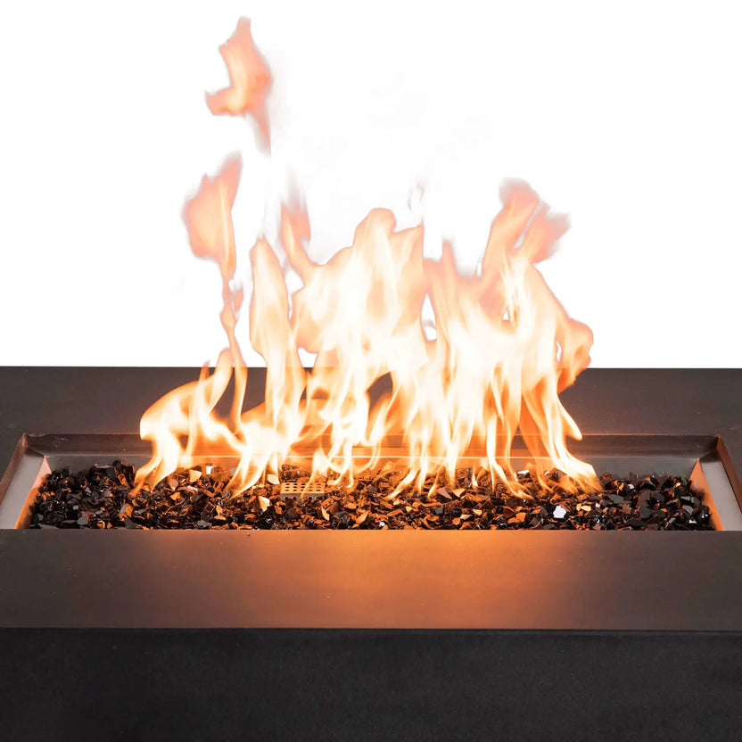 Close-up of the Flame Havens Refuge Series 56 x 30 x 15 inch rectangle natural gas fire pit with tall flames rising above reflective tempered black fire glass, highlighting the stainless steel burner system and modern black GFRC concrete surround.