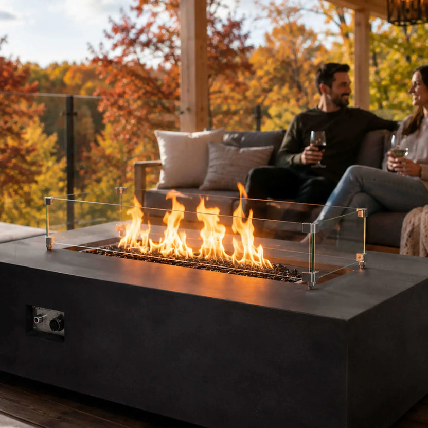 Flame Havens Refuge Series 56 x 30 x 15 inch rectangle natural gas black GFRC fire pit with tempered black fire glass and a tempered glass wind guard, showcased on a luxury outdoor patio where a couple enjoys the warm flames in a beautiful autumn setting.