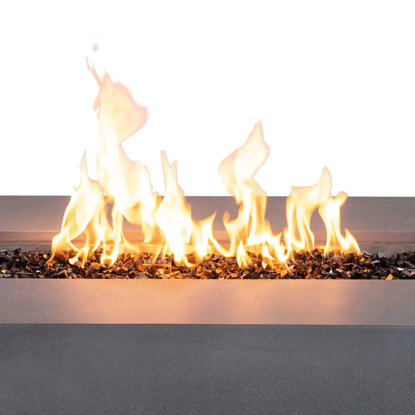 Close-up front view of the Flame Havens Refuge Series 56 x 30 x 15 inch rectangle natural gas gray GFRC fire pit burning with tall, vibrant flames over reflective tempered black fire glass. The image showcases the linear stainless steel burner system producing an even flame pattern across the full burner opening.