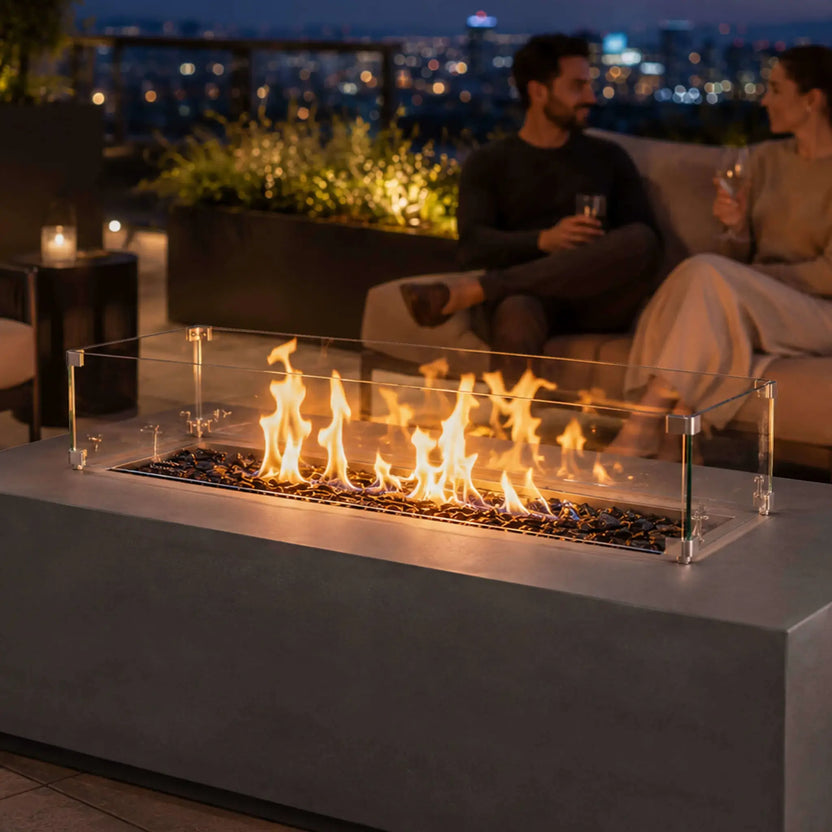 Lifestyle image of the Flame Havens Refuge Series 56 x 30 x 15 inch rectangle natural gas gray GFRC concrete fire pit with tempered glass wind guard installed. The fire pit glows on a luxury outdoor patio while two people relax nearby, showcasing reflective tempered black fire glass, vibrant flames, and an inviting setting for outdoor entertaining.