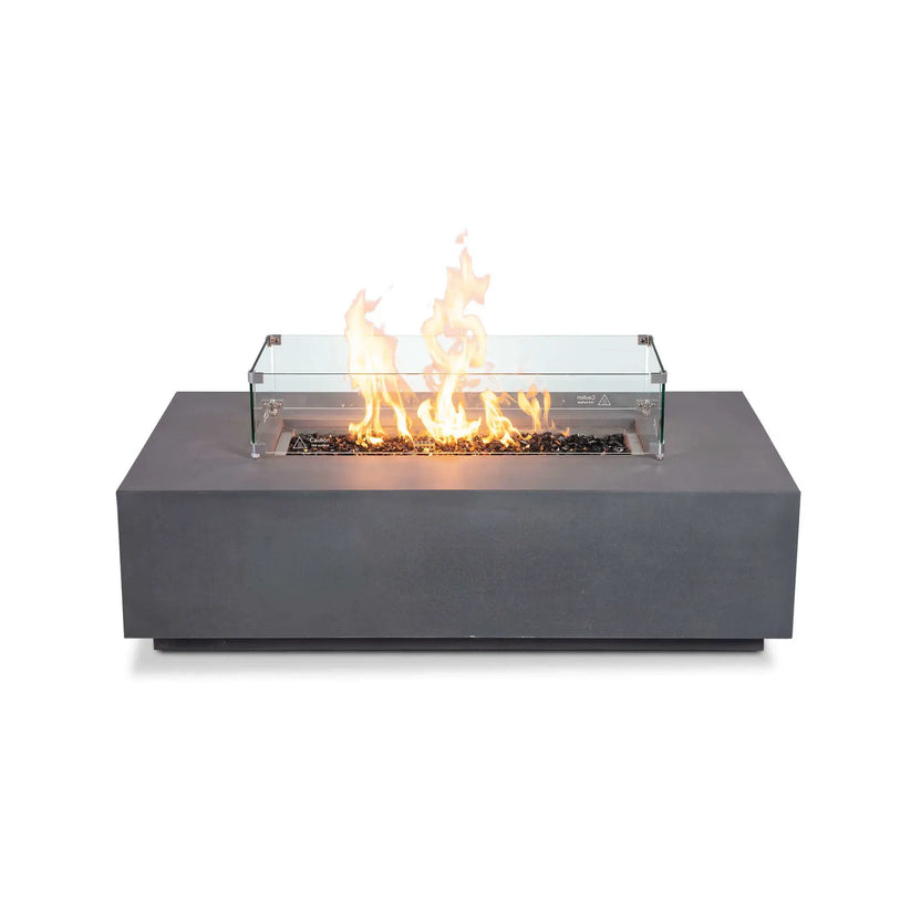 Straight-on view of the Flame Havens Refuge Series 56 x 30 x 15 inch rectangle natural gas gray GFRC concrete fire pit equipped with a tempered glass wind guard and reflective tempered black fire glass. The linear flame burns brightly behind the clear wind guard, demonstrating a premium outdoor fire feature designed for both style and enhanced flame protection.