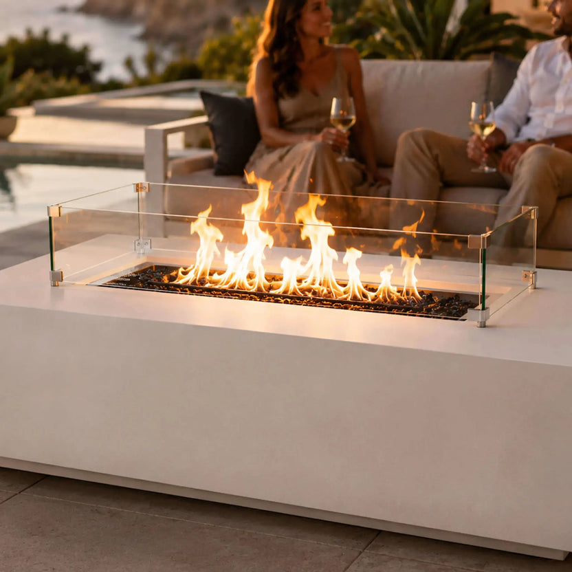 Lifestyle image of the Flame Havens Refuge Series 56 x 30 x 15 inch rectangle natural gas white GFRC concrete fire pit with tempered glass wind guard and reflective tempered black fire glass. A couple relaxes beside the illuminated fire feature overlooking a luxury pool and ocean at sunset, showcasing the fire pit as an elegant centerpiece for upscale outdoor entertaining.