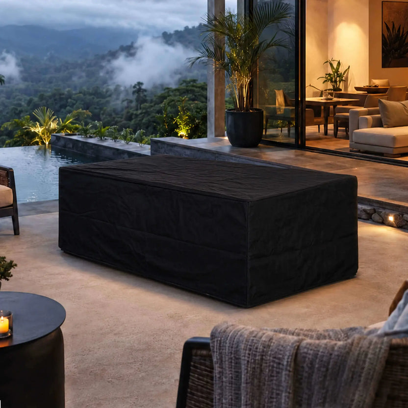 Black canvas fabric cover fitted over the Flame Havens Refuge Series 56 x 30 x 24-inch rectangular GFRC concrete propane gas fire pit on a luxury outdoor patio beside an infinity pool, with tropical mountain views and a warmly lit modern home in the background.